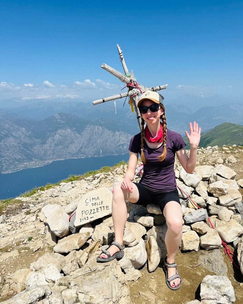 A hiker sits on the rocky summit of Cima delle Pozzette above Lake Garda and waves beside a wooden summit cross. Text reads "CIMA DELLE POZZETTE 2132 m".