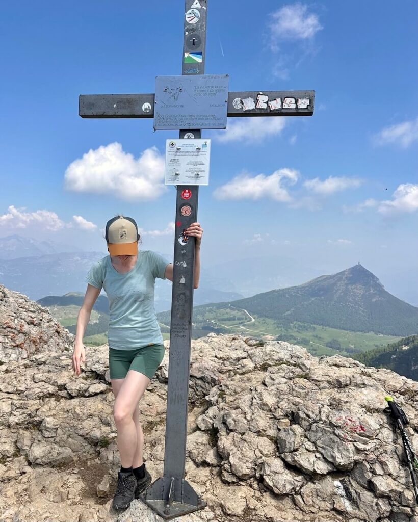 Hiker stands beside the summit cross on Monte Bondone Cornetto with rocky ground and green mountains behind her.