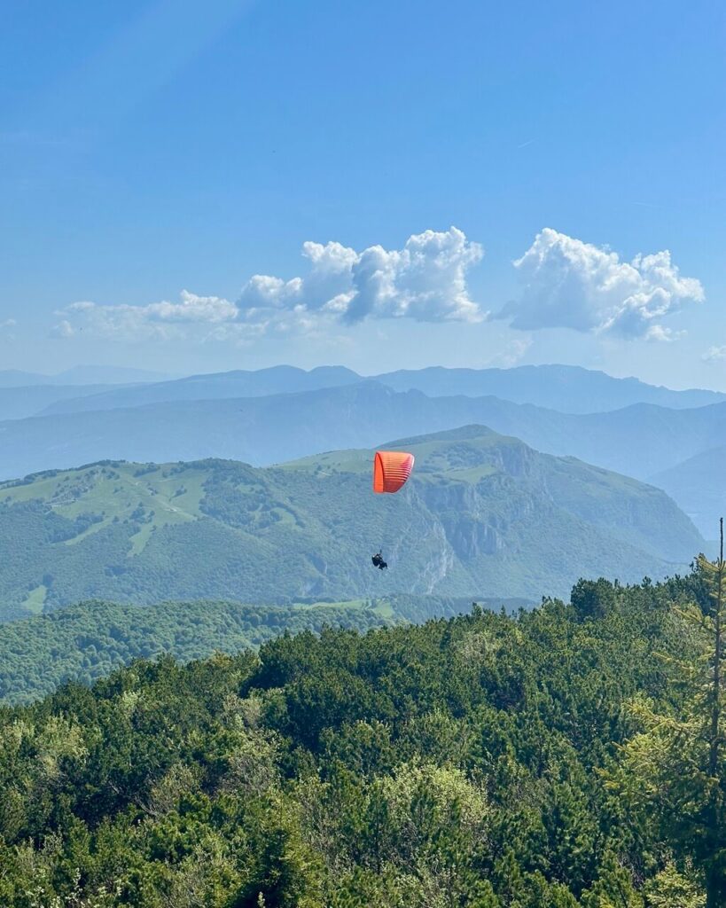 Orange paraglider floats above green mountain ridges and forest near Lago di Garda under a bright blue sky.