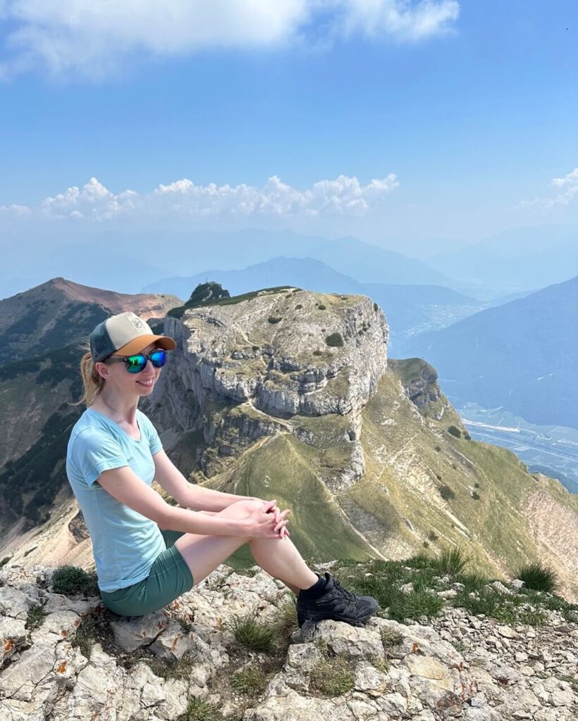 Hiker sits on pale summit rocks at Monte Bondone Cornetto with Dos d&rsquo;Abramo and hazy valleys behind her.