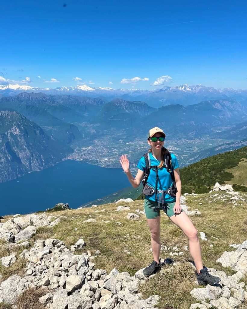 Hiker waves on a rocky ridge near Monte Altissimo di Nago with Lake Garda. Valley towns. Snow capped mountains. Wide alpine views behind her.