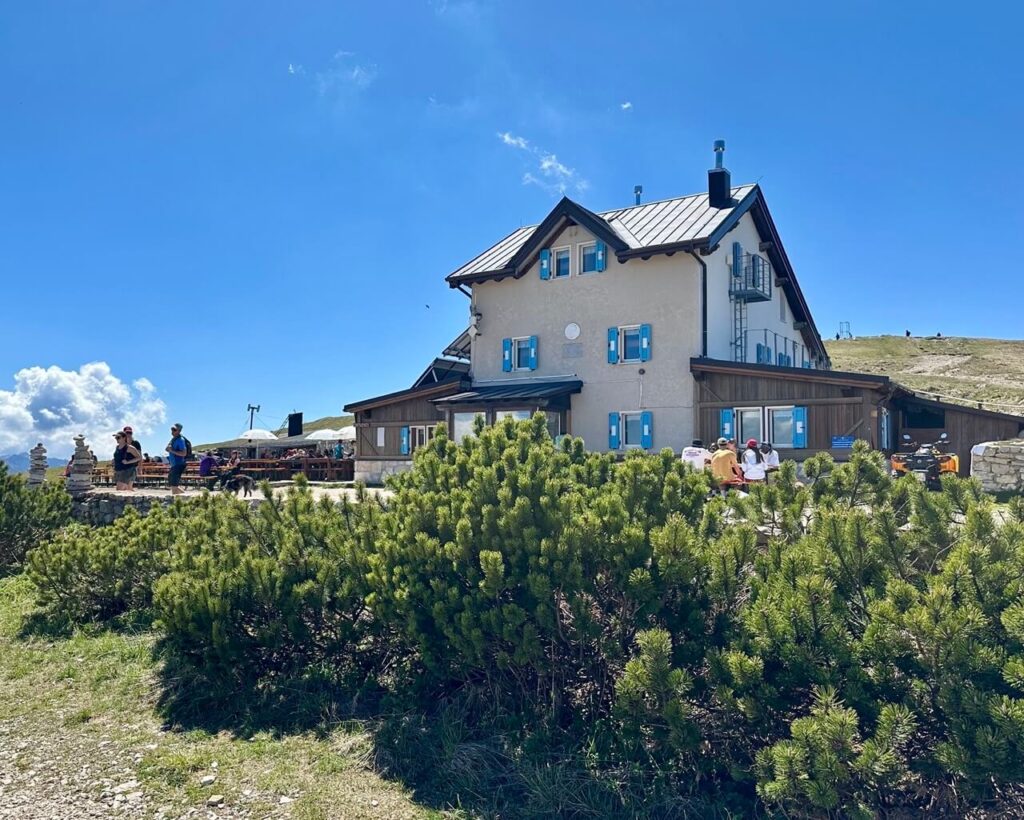 Mountain refuge with blue shutters beside a hiking trail near Monte Altissimo di Nago. Hikers gather on the sunny terrace behind low pine shrubs.