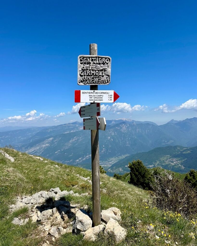 Wooden signpost on a mountain trail near Monte Altissimo di Nago. Text reads "SENTIERO DEI CIRMOLI" and "MALGA CAMPO 0.40" and "RIF. GRAZIANI 1.00" and "SAN GIACOMO 1.20".
