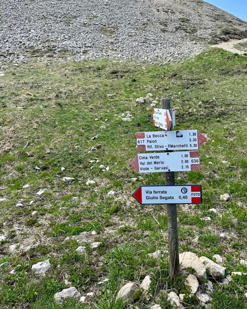 Hiking signpost below Monte Bondone Cornetto on a grassy rocky slope. Text reads &ldquo;La Becca 1.30&rdquo; and &ldquo;617 Palon 3.30&rdquo; and &ldquo;Rif. Stivo PMarchetti 5.30&rdquo; and &ldquo;Cima Verde 1.00&rdquo; and &ldquo;Val del Merlo 2.00&rdquo; and &ldquo;Viote Gervasi 2.15&rdquo; and &ldquo;Via ferrata Giulio Segata 0.40&rdquo;.