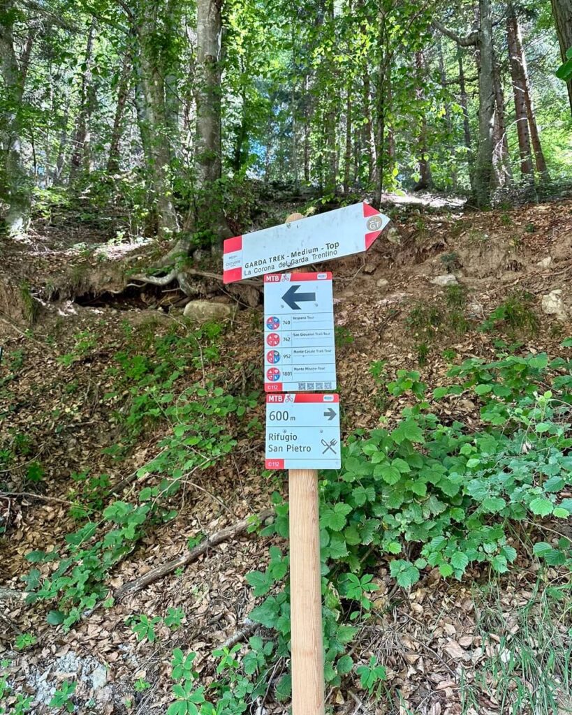 Trail signs in the forest point to Garda Trek and Rifugio San Pietro. Text reads &ldquo;GARDA TREK Medium Top. La Corona del Garda Trentino. MTB 9. 740 Vespana Tour. 742 San Giovanni Trail Tour. 952 Monte Casale Trail Tour. 1801 Monte Misone Tour. 600 m. Rifugio San Pietro&rdquo;.