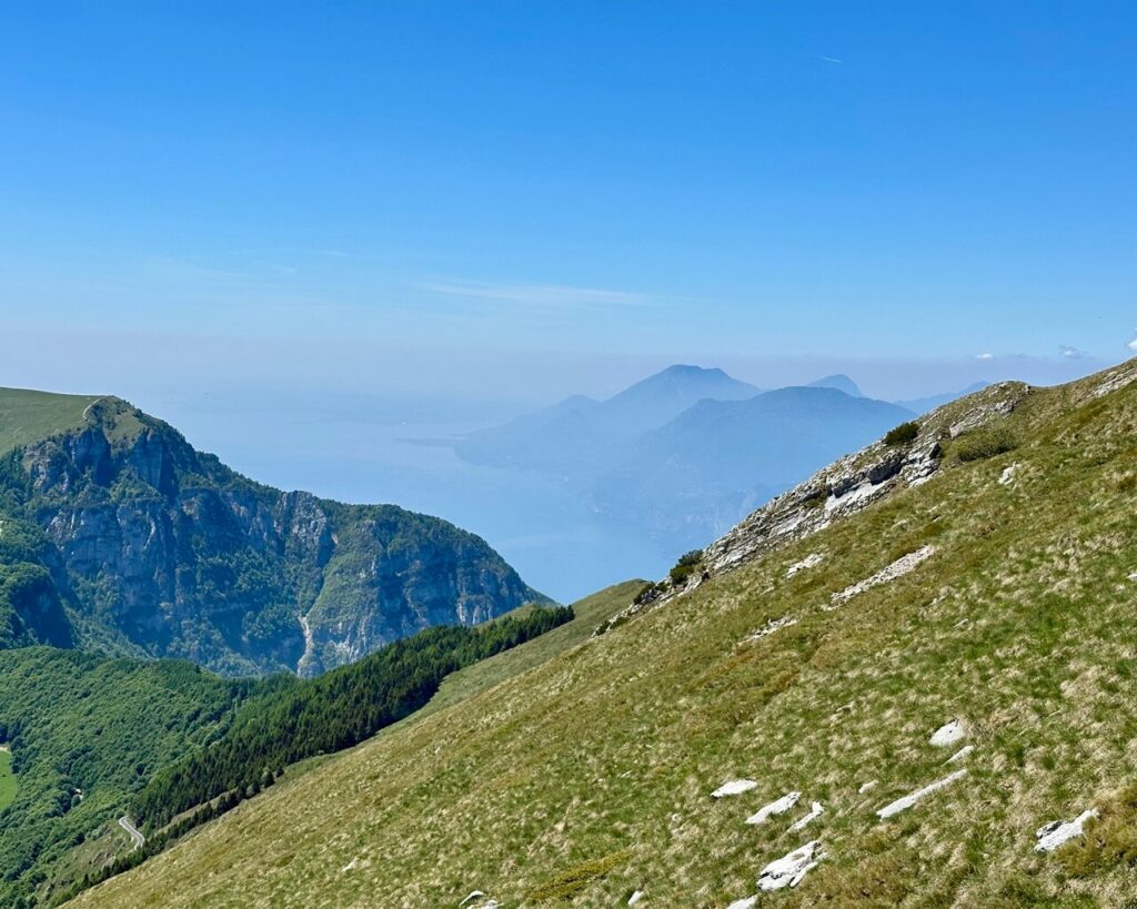 Sloping grassy mountainside near Monte Altissimo di Nago with pale rocks. Forested cliffs. Lake Garda fading into blue haze below.