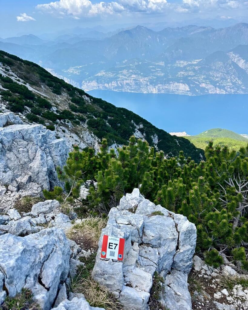 Rocky hiking trail above Lago di Garda with a red and white trail marker that reads "E7". Pine shrubs and pale limestone rocks frame the blue lake and distant mountains below.