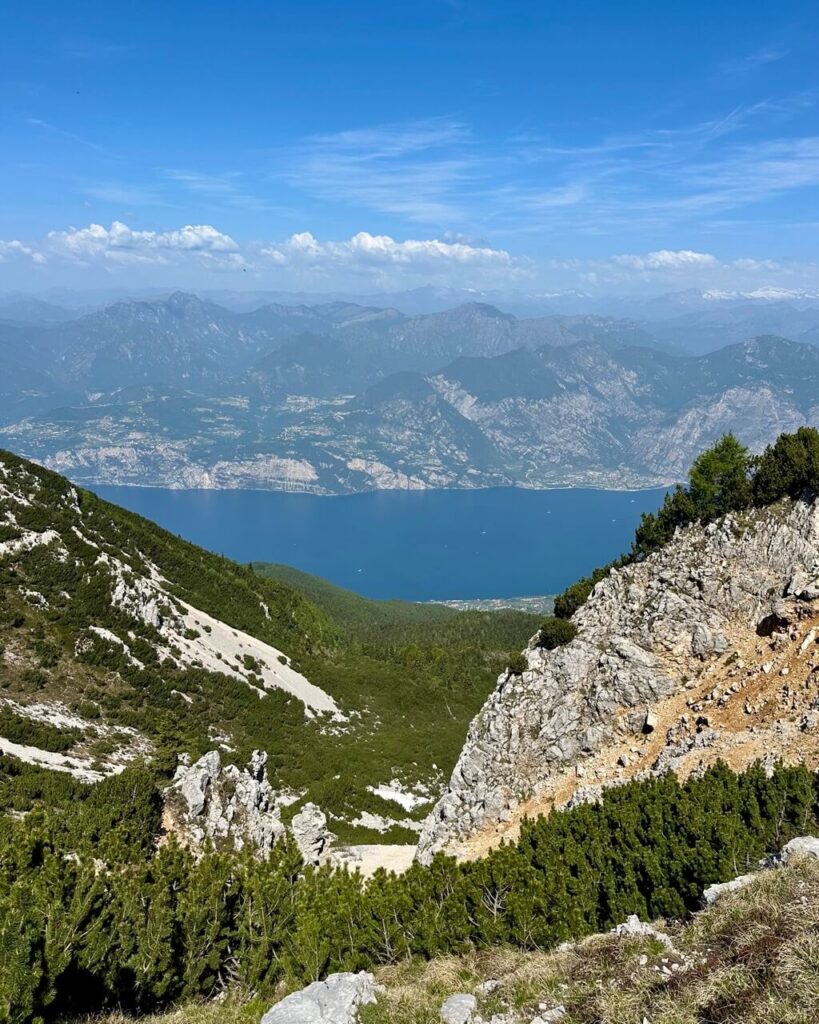 Wide Lake Garda hiking view from a rocky mountain trail with limestone cliffs. Green shrubs. Blue water far below. Hazy ridges across the lake.