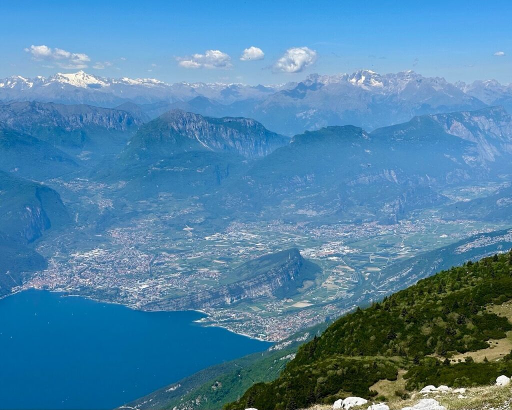 Expansive view from Monte Altissimo di Nago over northern Lake Garda Italy with blue water. Lakeside towns. Green valleys. Rocky mountains. Snowy peaks in the distance.