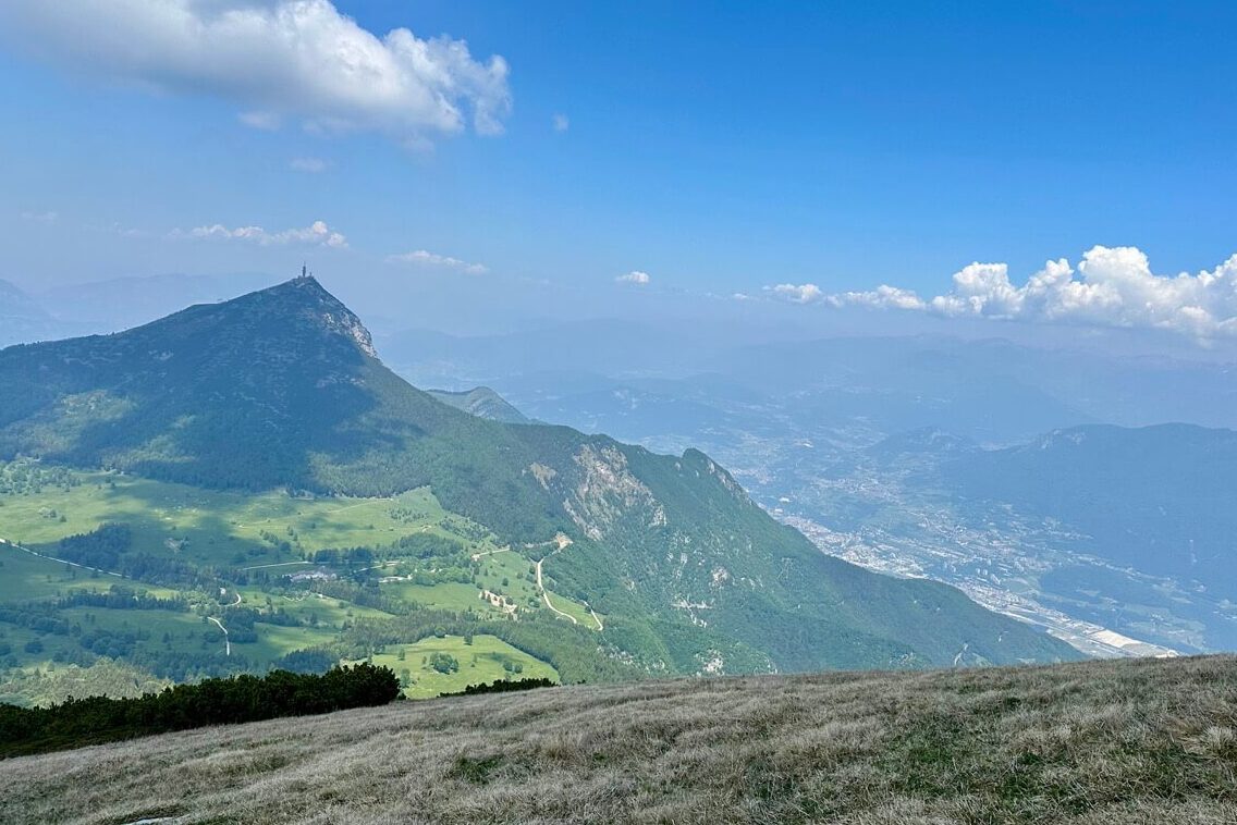 View from Cima Verde over green ridges and a pointed mountain with a summit structure above a hazy valley.
