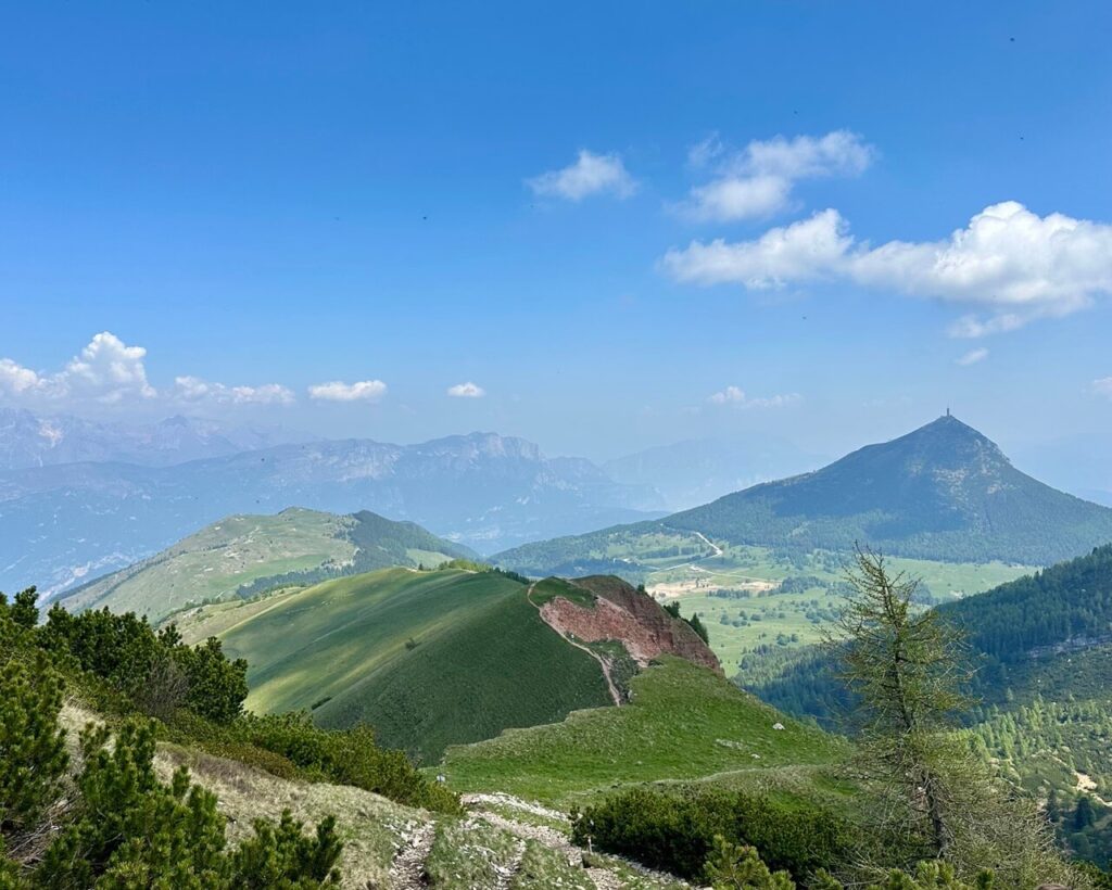 Panoramic view near Monte Bondone with rounded green ridges. A pointed mountain. Forested slopes. And hazy blue mountains beyond.